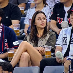 She is sitting casually in the stadium seats watching the baseball game.Legs crossed comfortably, occasionally adjusting posture naturally.Other spectators around her drinking beer, chatting, cheering, holding cheering sticks and mini portable fans. Plast