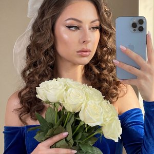 A girl is standing indoors in front of a neutral background, holding a large bouquet of white roses with her left hand. Her left hand is positioned gently around the stems and leaves of the flowers, fingers softly resting on the green foliage. Her right h