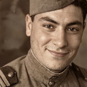 Реалистично анимируй фотографию с естественными движениями человека. Человек мягко улыбается, затем начинает приветливо махать рукой в сторону камеры. Живая мимика, естественное моргание, лёгкие движения головы и тела. Сохрани исходную внешность, одежду, 