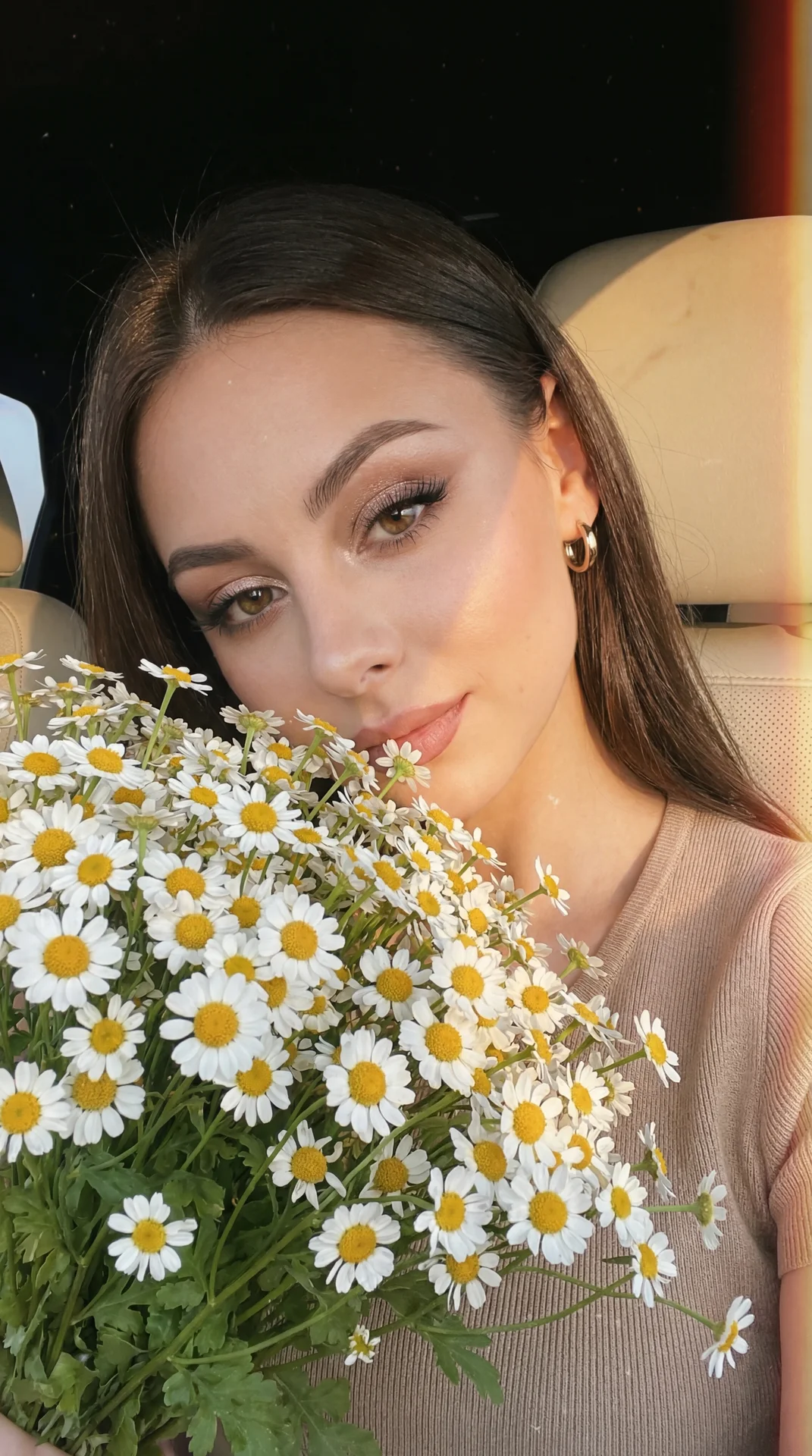 A girl takes a selfie in close-up, slightly tilted forward-on the right at eye level (85 mm, f/1.8), in the car at the window, lighting is a sunset fan, sunset, rays fall beautifully on objects; a bouquet of white daisies is held up to her face, flowers d
