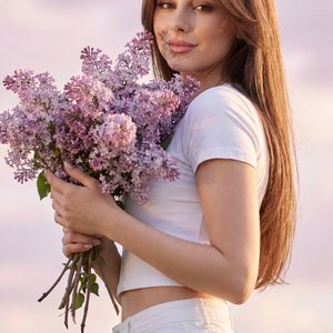 Professional portrait, medium shot (waist-up), slight low angle, three-quarter body turn; outdoor at golden hour under pastel pink-lilac clouds. Female model holds a large lilac bouquet at chest height—front hand (closer to camera) cradles the stems from 