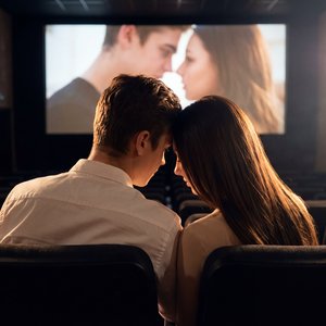СТРОГО сохранить лицо, кожу, возраст, черты и пропорции 1:1 без любых изменений. Кинотеатр: темный зал с рядами кресел, большой экран впереди; на экране — крупный кадр пары; в кадре перед камерой сидит пара: мужчина в белой рубашке и женщина в светлом топ