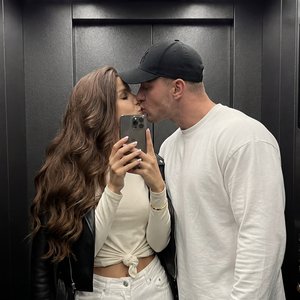 Фотореалистичное lifestyle‑фото как из Pinterest, снято на заднюю основную камеру смартфона (1x). Вертикальный кадр 9:16. Это зеркальное фото в лифте: пара стоит близко к зеркалу, девушка держит смартфон в руке и снимает отражение. Натуральная цветопереда