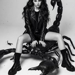 A girl, full-body shot, slight low-angle, professional DSLR photography. Slender figure. Posed sitting astride a massive, highly detailed black scorpion sculpture. Her legs are spread apart, knees bent, with her left foot planted firmly on the studio floo