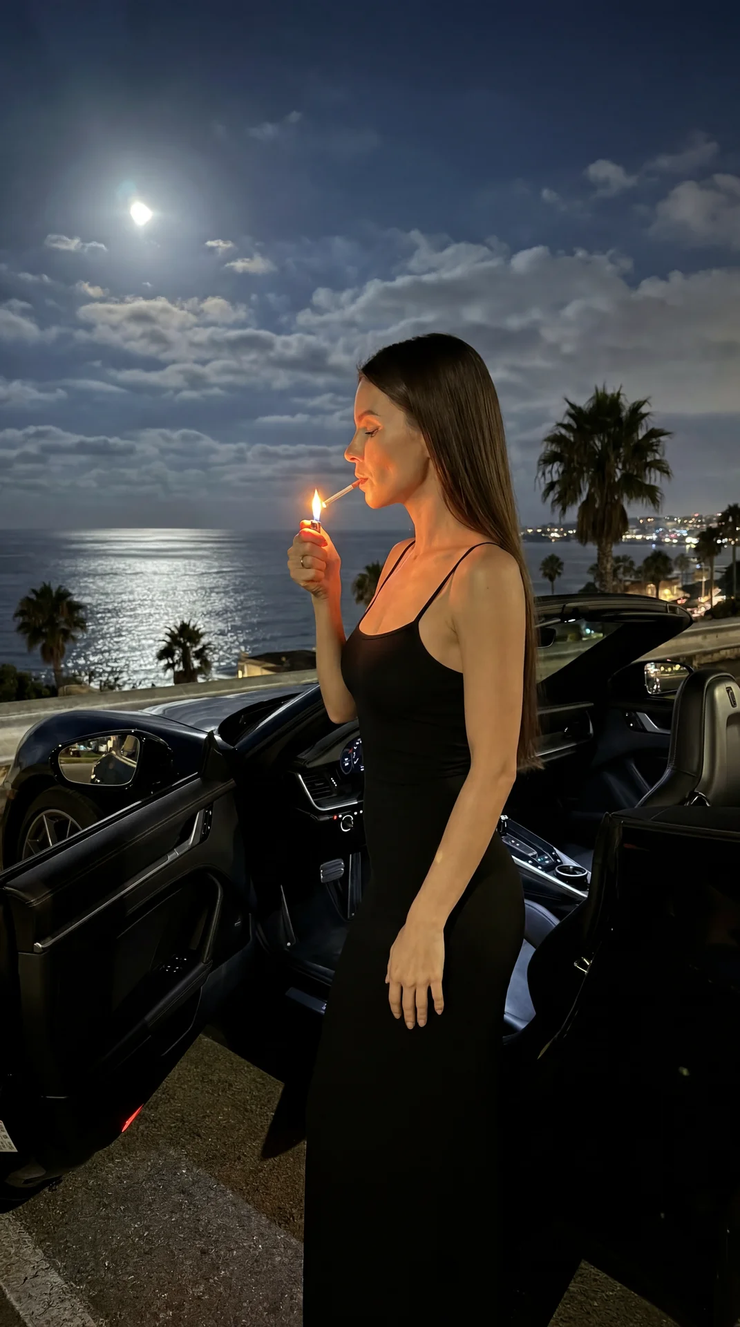 Shot on iPhone 15 Pro, aspect ratio 3:4, no flash, natural night photo. A woman stands next to the open driver’s door of the latest generation black convertible Porsche 911, parked at night at the coastal observation dechange. She’s in profile with her fa