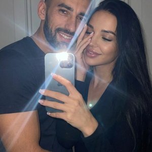 Зеркальное селфи молодой пары, снятое под немного высоким углом. мужчина располагается слева, обхватив рукой девушку, находящуюся справа. Оба мягко улыбаются, глаза парня видно , у девушки закрыты или частично затенены.Мужчина он одет в темную футболку . 