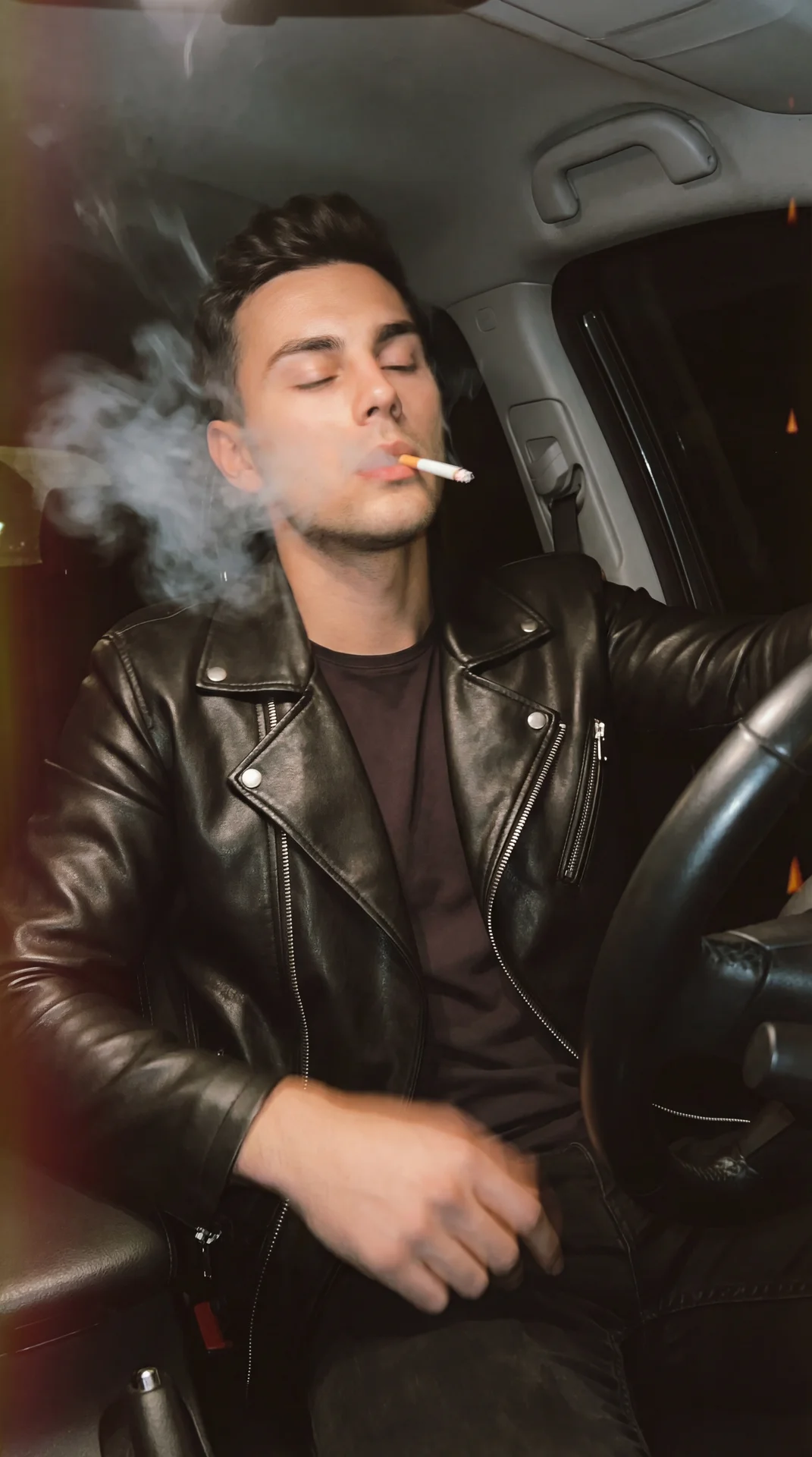 A men sitting in the driver’s seat of a car at night, medium close-up, shot from inside the cabin at a slight low angle with the steering wheel in the foreground framing the right side; head slightly tilted back, eyes gently closed, mouth slightly open ho