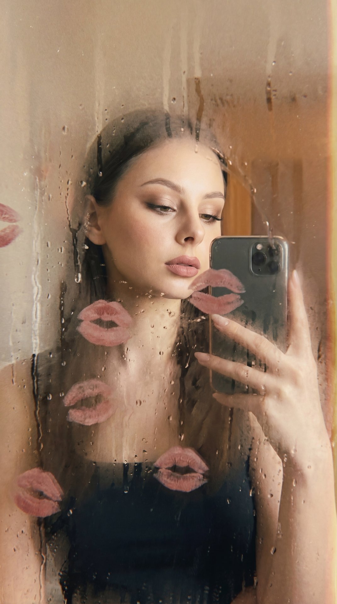 A girl is standing behind a fogged glass surface covered with water droplets and streaks, which partially obscure her figure and face, creating a soft, blurred effect. She holds a smartphone in her right hand raised to a mid-chest level, capturing a mirro