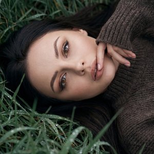 Гиперреалистичное фото 9:16, как живая фотография; сохранить прическу и черты лица без изменений; вертикальный крупный план, ракурс сверху с легким наклоном, камера смотрит почти перпендикулярно вниз; девушка лежит на спине в густой влажной траве с каплям