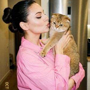 A girl with slicked‑back hair gathered in two voluminous buns, dressed in a spacious bright pink cotton poplin button-down shirt, stands in a room near a stainless steel refrigerator; the body is three-quarters turned to the camera to the right, a straigh