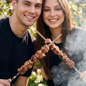 Cinematic portrait of a happy young couple at a sunny dacha backyard, looking directly at the camera and smiling broadly. They are playfully raising metal skewers with hot, juicy shashlik like a toast. Smoke softly rising in the background, blooming sprin