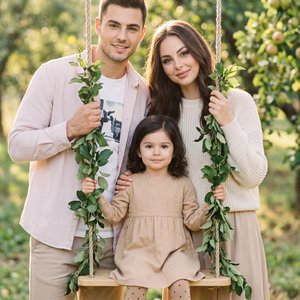 Professional family portrait in a sunlit orchard, vertical medium-wide shot, eye-level camera, 85mm lens, f/2.0, soft backlight and creamy bokeh, warm pastel color grading, crisp subjects with gentle rim light; a man (left) and a woman (right) flank a woo