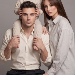 Professional studio family portrait, straight-on eye-level medium shot on a short‑telephoto lens (minimal distortion, crisp detail), neutral gray seamless backdrop, soft diffused key from front‑left with gentle right fill, even skin tones, minimal shadows