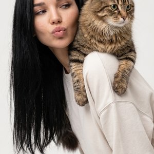 Студийный fashion-портрет на светлом фоне. Девушка слегка наклонена вперёд, корпус в полуповороте. На её плече спокойно лежит кот мэйнкун, передними лапами обнимая плечо. Девушка делает лёгкий «поцелуй» губами в сторону, жест игривый, момент живой и естес