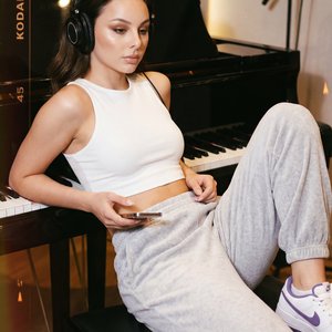 A girl is captured in a relaxed, candid medium shot leaning back on a black piano bench in a recording studio. The camera angle is slightly high and tilted, emphasizing her casual, comfortable pose and the environment around her. She reclines on the bench
