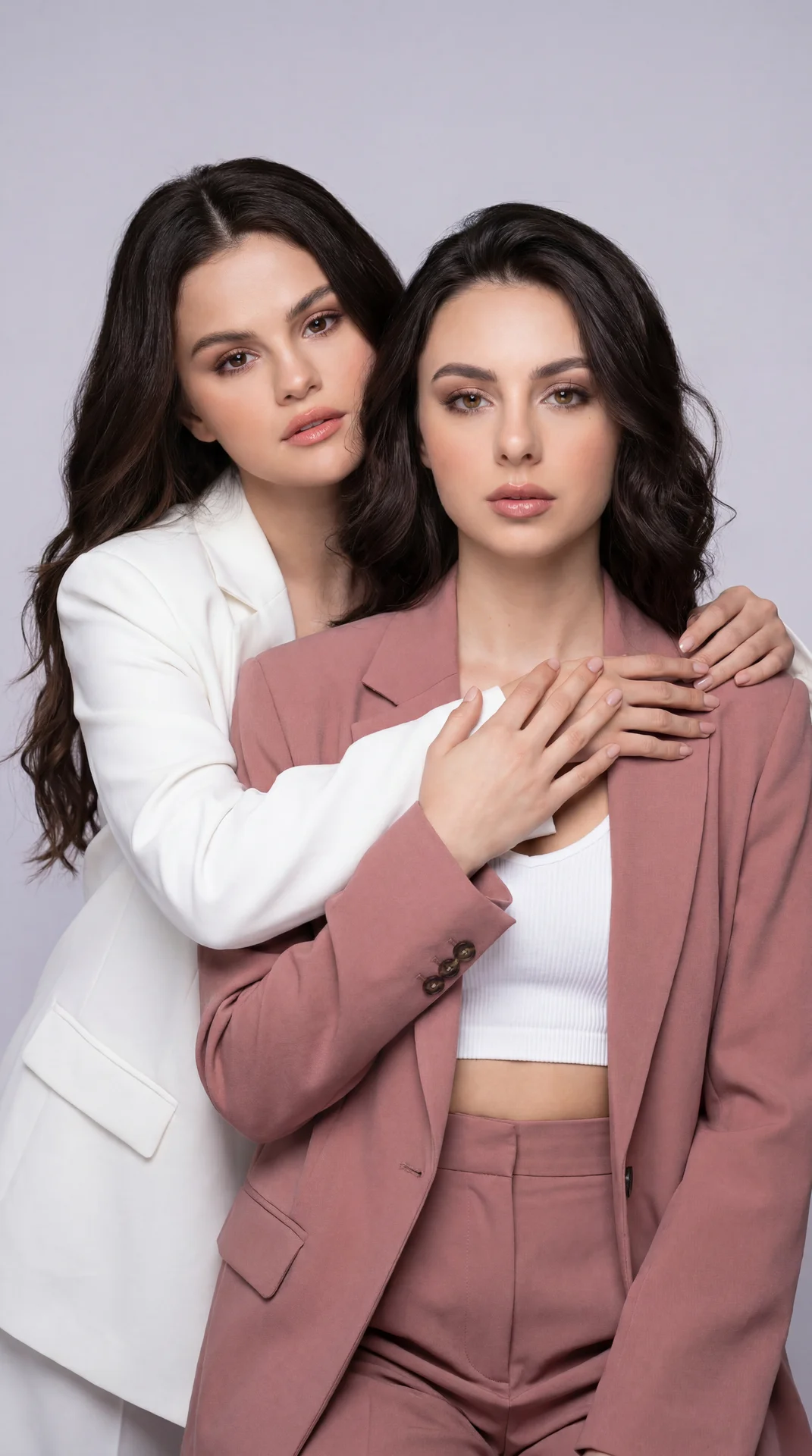 Soft pastel studio fashion duo, two adult women, front model in dusty rose oversized suit (matte crepe/wool-blend blazer and high‑waist tailored trousers) with a white ribbed crop top; rear model in a clean white oversized blazer (smooth matte fabric). Po