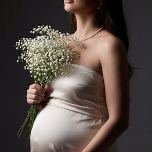 Professional studio portrait, medium shot, three-quarter profile of a pregnant woman standing; eyes gently closed, head subtly tilted upward toward camera-left, serene calm expression, lips softly closed; one hand cradles the underside of the belly (palm 