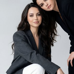 Two female models in a minimalist studio, medium three-quarter portrait, eye-level straight-on angle, professional camera look (85mm, f/2.8), soft diffused studio light, cool-neutral grading, subtle film grain, gentle vignetting. Front model crouched with