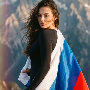 A girl, medium waist-up shot, over-the-shoulder camera angle, professional DSLR photography. Athletic slender figure. Standing with her back towards the background, torso gracefully twisted backward over her left shoulder to face the lens. Left arm bent d