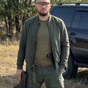 Высококачественное фото молодого мужчины в стиле military. На нем приталенная темно-зеленая (болотного цвета) куртка-бомбер без воротника, надетая поверх оливковой базовой футболки, и широкие темно-зеленые тактические штаны с карманами, на правом бедре та