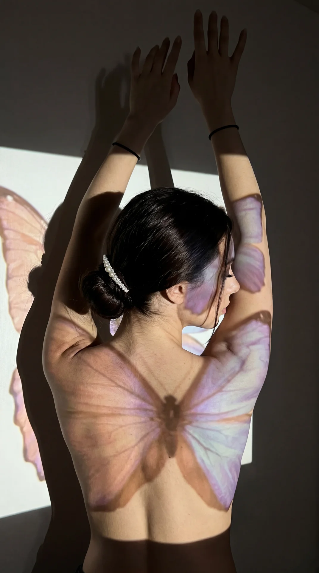 A moody, vertical 9:16 photo taken on an iPhone in a dimly lit room at night — likely a bedroom or studio space with plain walls and minimal decor. The subject is a young woman seen from behind, standing close to a wall where projected light casts dramati