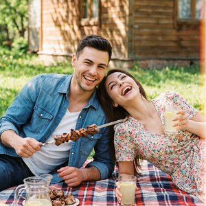Top-down angle cinematic photography, a young couple relaxing on a cozy plaid blanket on green grass near a wooden dacha house. They are eating grilled shashlik from metal skewers, drinking lemonade, and laughing hard. Casual spring outfits, bright sunny 