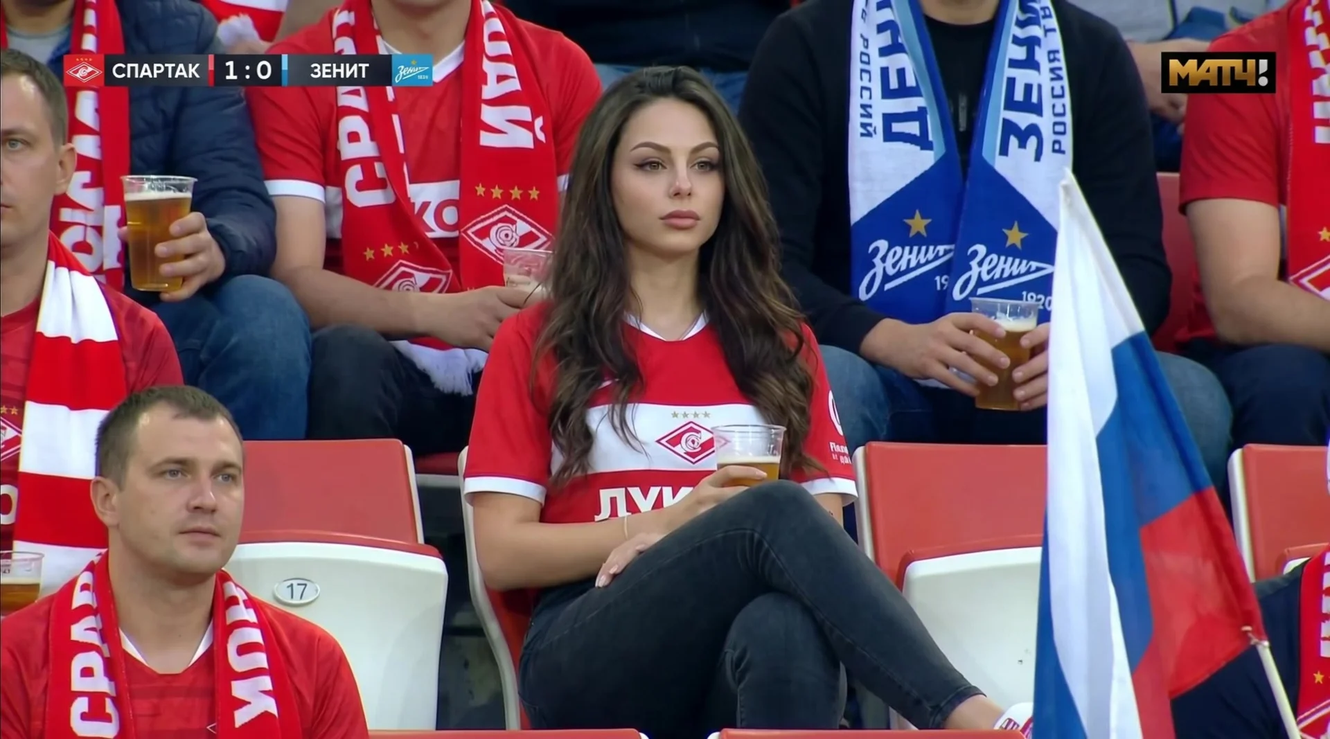 A realistic sports broadcast-style photo shoot taken in the stands of a stadium during a professional football match in Russia. The attached image shows a woman sitting in the stands. She has delicate features and a surprised yet focused expression, looki