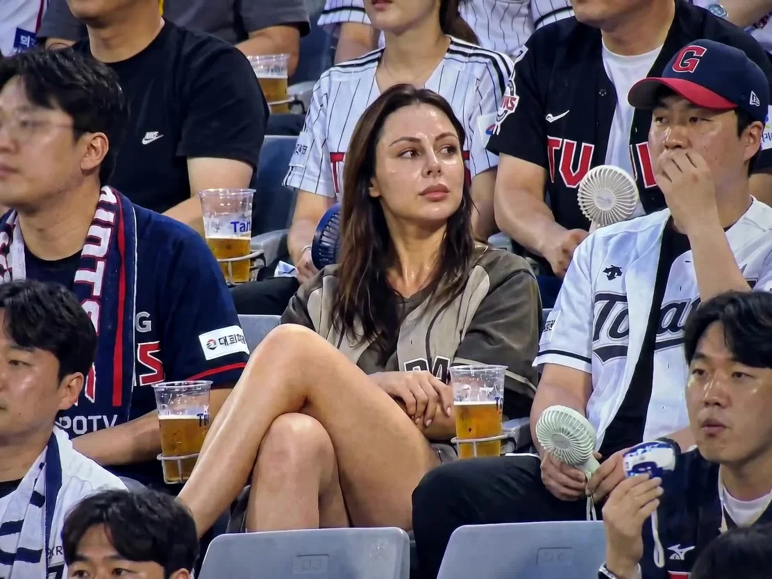 She is sitting casually in the stadium seats watching the baseball game.Legs crossed comfortably, occasionally adjusting posture naturally.Other spectators around her drinking beer, chatting, cheering, holding cheering sticks and mini portable fans. Plast