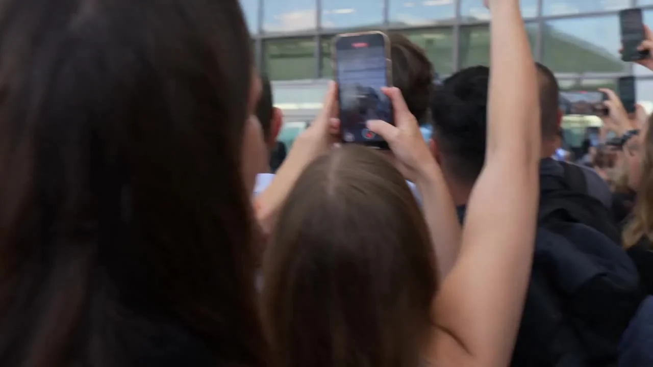 Style: Ultra realistic mass celebrity entry scene. Single continuous shot. Handheld camera from crowd perspective. Natural micro-shake. No cuts. Documentary realism.
Audio: Only natural environment sound — loud crowd cheering, people shouting, rapid camer