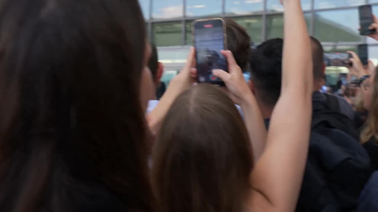 Style: Ultra realistic mass celebrity entry scene. Single continuous shot. Handheld camera from crowd perspective. Natural micro-shake. No cuts. Documentary realism.
Audio: Only natural environment sound — loud crowd cheering, people shouting, rapid camer