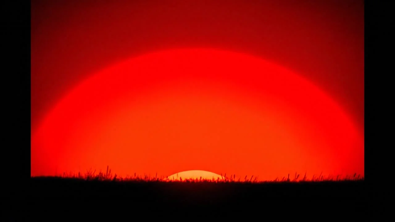 Cinematic slow sunrise animation based on the reference image. A large deep red sun slowly rises from behind a dark grassy horizon. At the beginning of the video the sun is already about 10% above the horizon, and during the 23-second shot it gradually ri