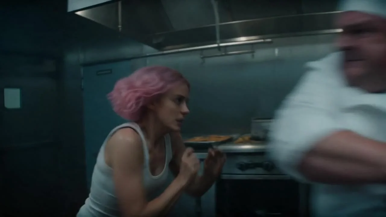 A young woman @image_1 with pink hair in a white tank top and jeans fights a heavyset chef in a dimly lit commercial kitchen. Dynamic handheld camera tracks the intense close-quarters action as she dodges between stainless steel counters and baking racks.